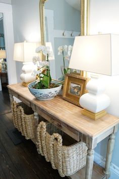 a wooden table topped with a bowl of flowers next to a mirror and lamp on top of it