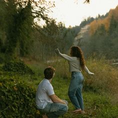 two people are sitting on the ground in front of some bushes and trees, one person is pointing at something