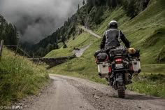 a man riding on the back of a motorcycle down a dirt road next to a lush green hillside