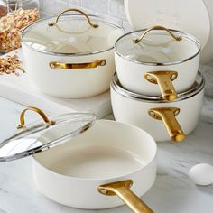 white pots and pans with gold handles sitting on a marble countertop next to other dishes