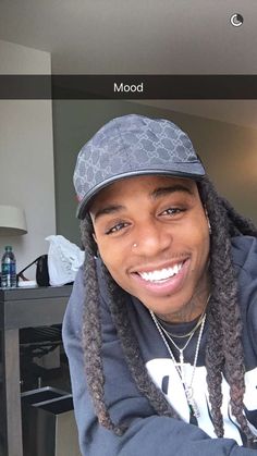 a young man with dreadlocks and a hat smiling at the camera while sitting in front of a table