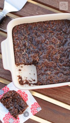 a brownie in a white baking dish on a wooden table next to a fork and napkin