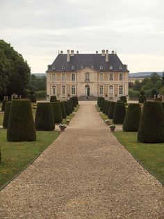 a large house surrounded by hedges in front of a gravel path with potted plants on either side