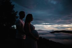 a pregnant couple standing on top of a mountain at night with clouds in the background