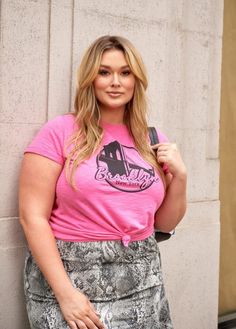 a woman in a pink shirt is leaning against a wall and posing for the camera