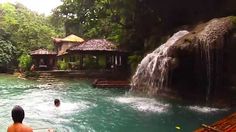 two people are in the water near a waterfall and some huts with thatched roofs