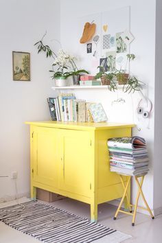 a yellow cabinet with books and plants on top in a room that has white walls