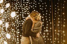 a man and woman embracing each other in front of a wall covered with white lights