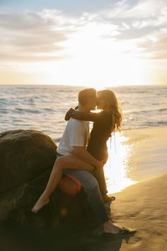 two people sitting on top of a large rock near the ocean with sunset in the background