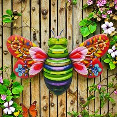 a colorful insect sculpture sitting on top of a wooden fence surrounded by flowers and butterflies