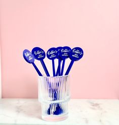 six blue plastic spoons in a clear glass on a marble countertop against a pink wall