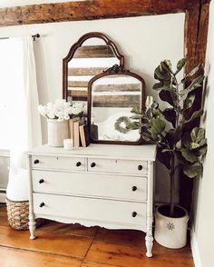 a white dresser sitting next to a window with a mirror and potted plant on top