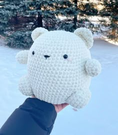 a hand holding up a crocheted stuffed animal in the snow with pine trees in the background