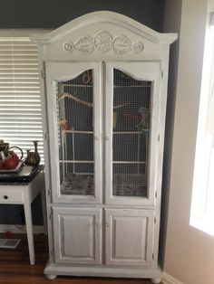 an old white china cabinet with birdcages on the top and bottom doors, next to a window