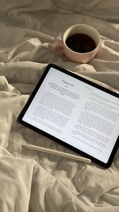 a tablet and pen sitting on top of a bed next to a cup of coffee