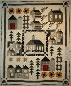 a quilted wall hanging with houses and sunflowers in the center, on display