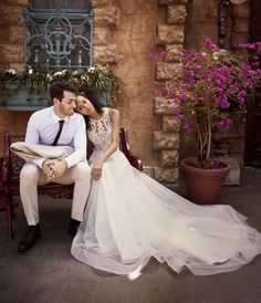 a bride and groom sitting on a bench in front of a building with purple flowers