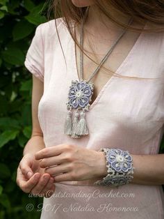 a woman wearing a necklace with tassels and beads on her wrist is standing in front of some bushes