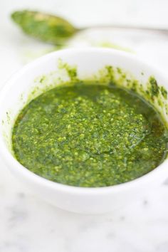 a white bowl filled with green pesto on top of a marble counter next to two spoons