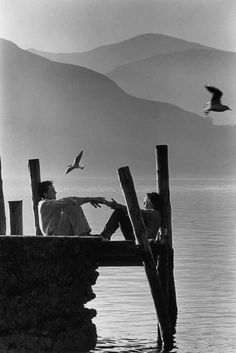 black and white photograph of two people sitting on a dock with seagulls flying over the water