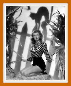 a black and white photo of a woman sitting in front of corn stalks with a cat shadow on the wall behind her