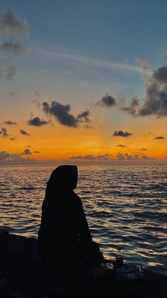 a person sitting on the shore watching the sun go down over the ocean with clouds in the sky