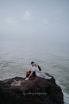 a man and woman sitting on top of a rock near the ocean with their arms around each other