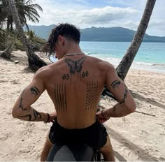 a man with tattoos on his back sitting on a beach next to the ocean and palm trees