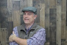 a man wearing a hat and vest standing in front of wooden wall with his arms crossed