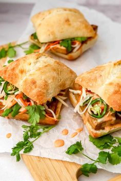 three sandwiches with meat and veggies are on wax paper next to a cutting board