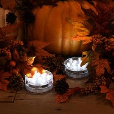 two lighted candles surrounded by autumn leaves and pine cones on a wooden table with a pumpkin in the background