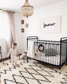 a baby standing next to a black crib in a white room with a chandelier hanging from the ceiling