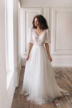 a woman in a white dress is standing near a window and looking off to the side