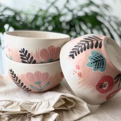 two pink and blue bowls sitting on top of a white table cloth next to a potted plant