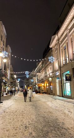 two people walking down a snowy street in the middle of town at night with christmas lights strung above