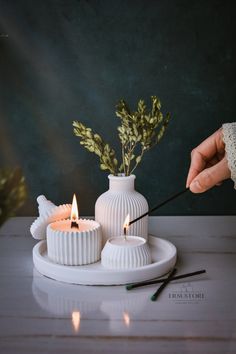 a person lighting candles on top of a white plate next to a plant in a vase