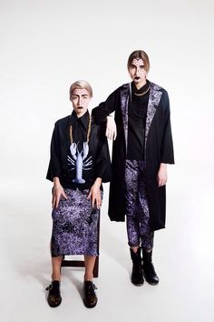 two people standing next to each other in front of a white background wearing black and purple clothing