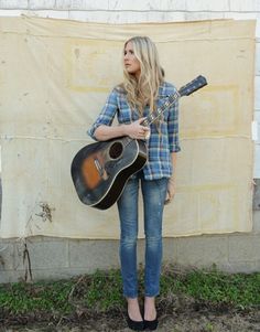 a woman holding a guitar standing in front of a wall with a tarp behind her