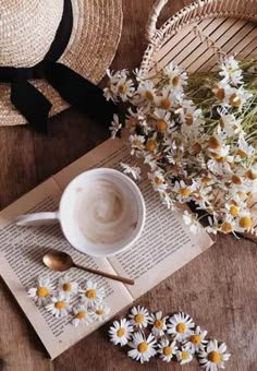 a cup of coffee sitting on top of an open book next to daisies and a straw hat