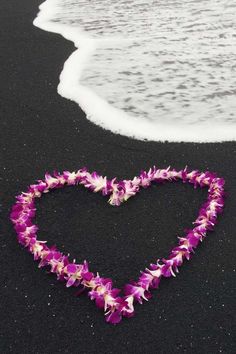 purple flowers arranged in the shape of a heart on a black sand beach with waves coming in