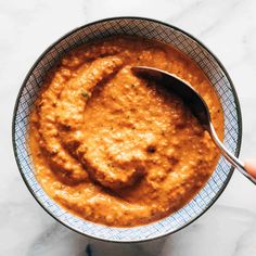 a hand holding a spoon over a bowl of red sauce on a marble counter top