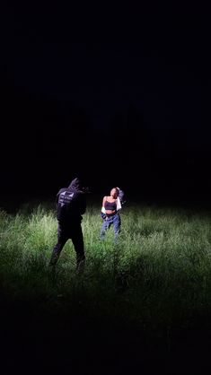 two people standing in the grass at night, one is holding a camera and the other is wearing a backpack