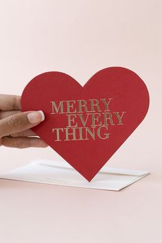 someone holding up a heart shaped card with the words merry everything on it in gold foil