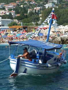 a man in a blue and white boat on the water near a crowded beach with umbrellas