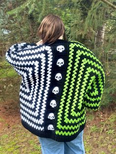 a woman wearing a black and green knitted sweater with skulls on the back, standing in front of trees