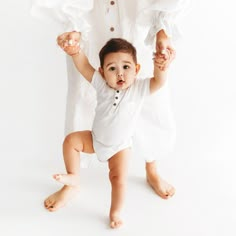 a baby standing in front of a white backdrop holding onto her mother's hands