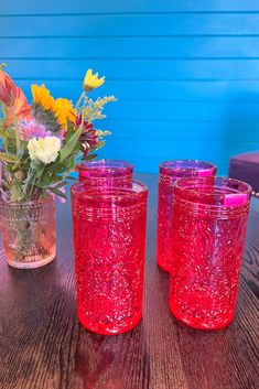 three red glasses sitting on top of a table next to a vase filled with flowers