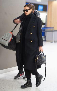 a woman in black coat and sunglasses carrying two handbags while walking through an airport