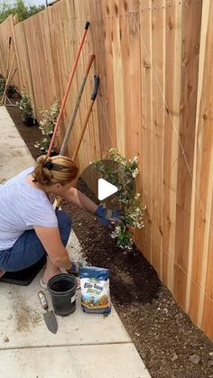 a woman kneeling down next to a wooden fence with gardening tools in her hand and potted plants on the ground