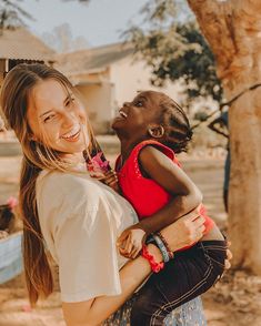 a woman holding a child in her arms and smiling at the camera while standing next to a tree